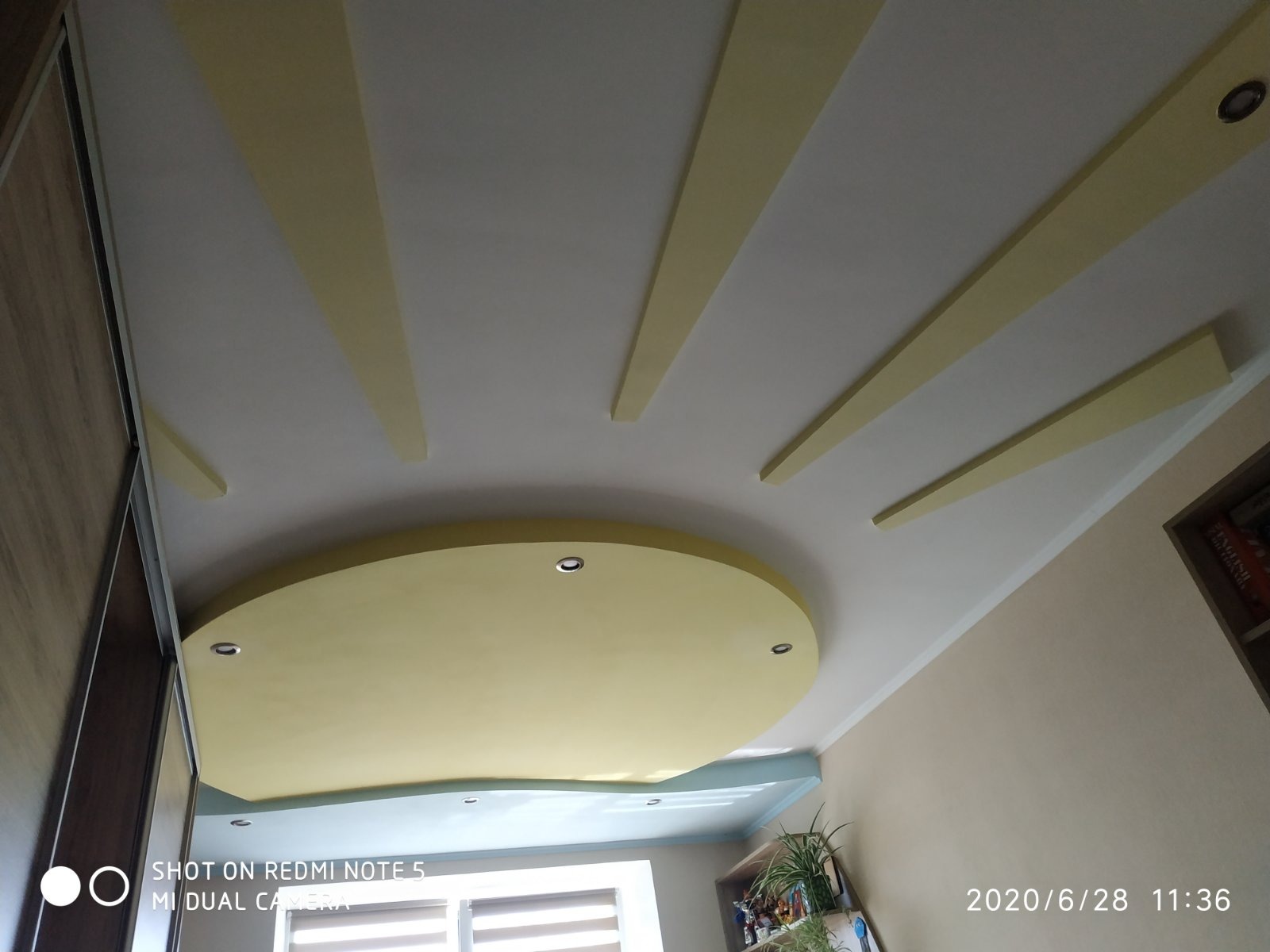 Żółto-biały sufit podwieszany z oświetleniem punktowym i dekoracyjnymi listwami. Ujęcie z dołu, widoczny fragment szafy i okna.
