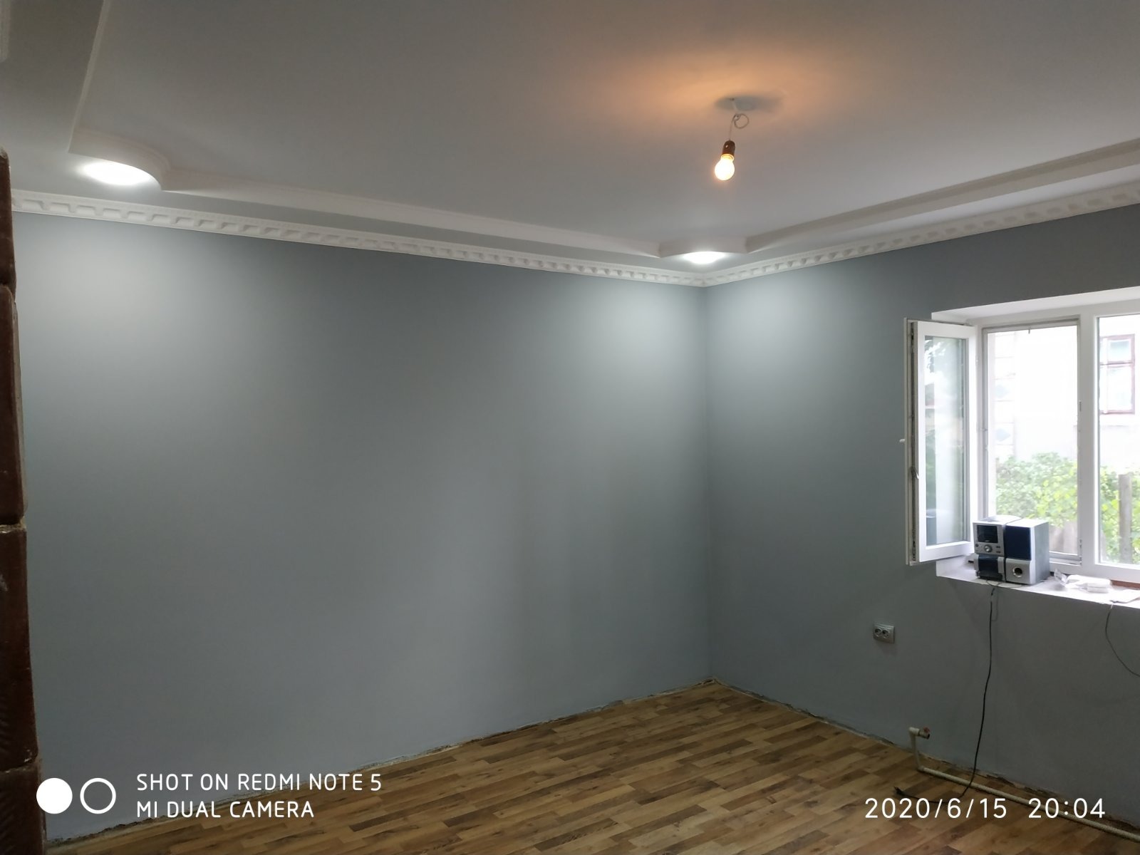 Pokój po malowaniu ścian na szaro, z drewnianą podłogą i białym sufitem z dekoracyjną listwą. Widoczne okno z parapetem i sprzętem audio.