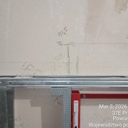 Bulba - Metalowy stelaż pod zabudowę, widoczne profile i oznaczenia na ścianie. Elementy instalacji sanitarnej w trakcie montażu. Ściana z surowym wykończeniem.