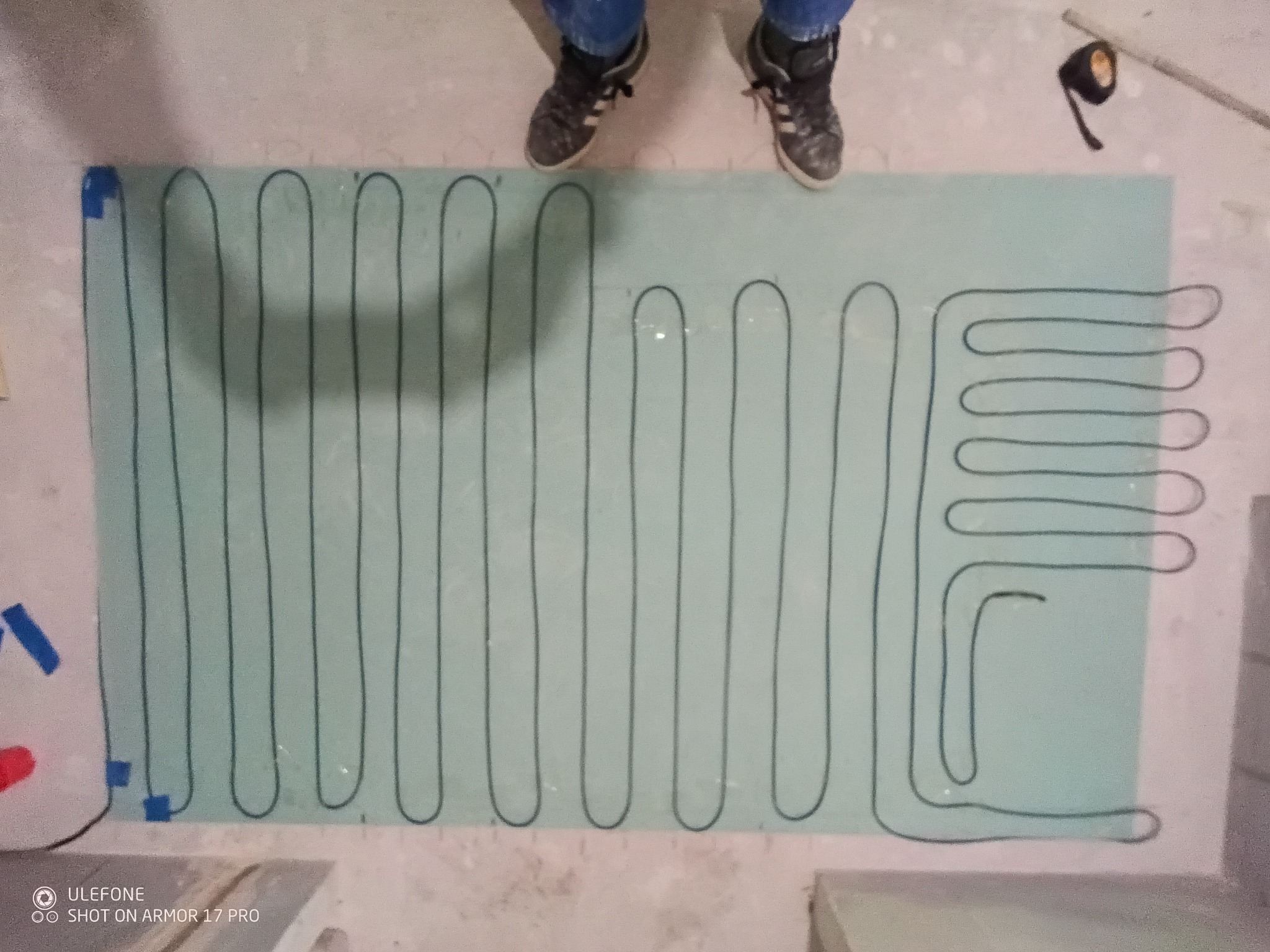 Ogrzewanie podłogowe: czarny przewód ułożony na jasnej płycie izolacyjnej, widok z góry, stopy w butach na płycie, taśma izolacyjna i fragment ściany w tle.