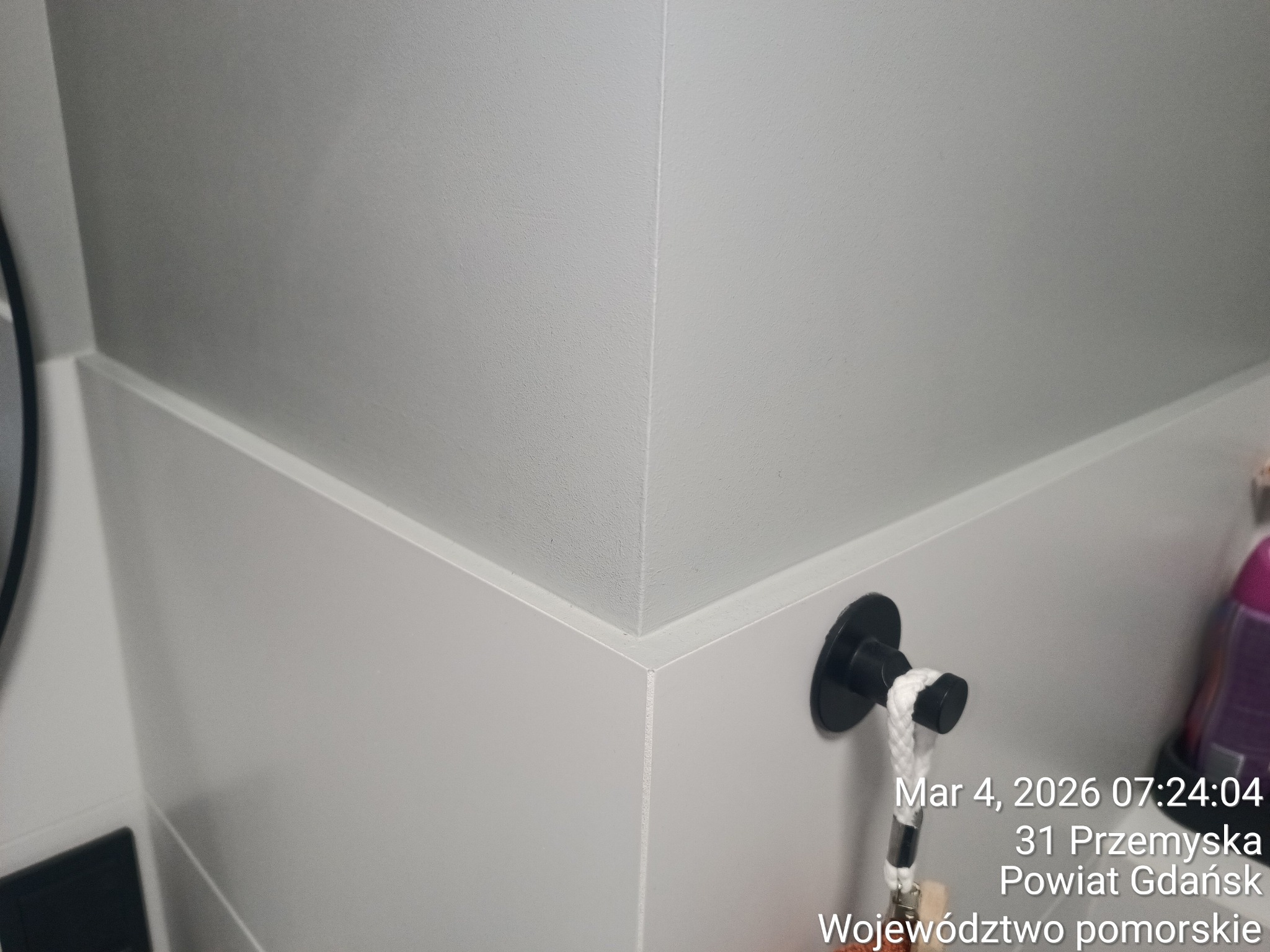 Szare płytki ścienne w łazience, z widocznym czarnym haczykiem na ręcznik i fragmentem butelki. Minimalistyczny design, gładkie powierzchnie, nowoczesne wykończenie.