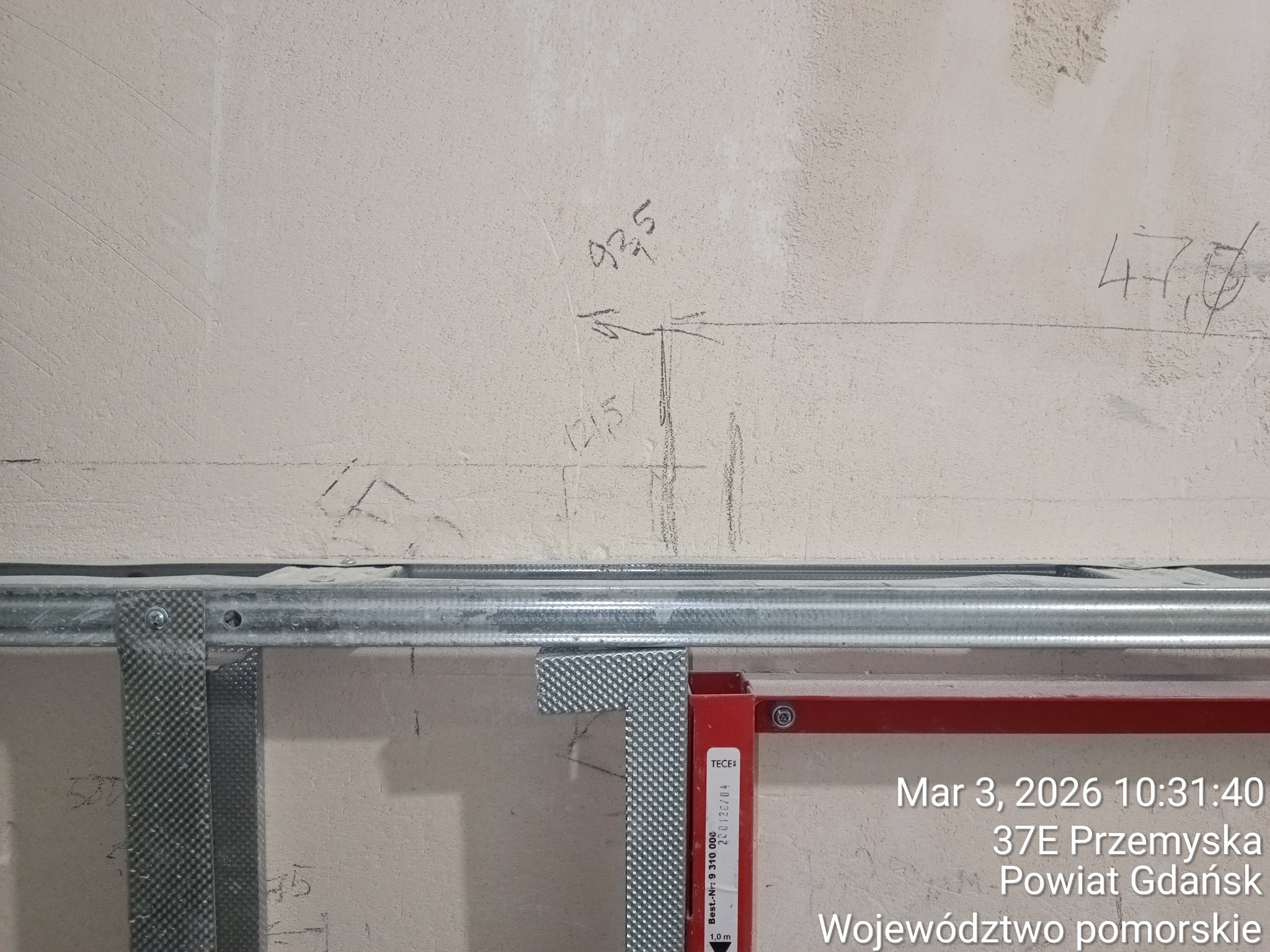 Metalowy stelaż pod zabudowę, widoczne profile i oznaczenia na ścianie. Elementy instalacji sanitarnej w trakcie montażu. Ściana z surowym wykończeniem.