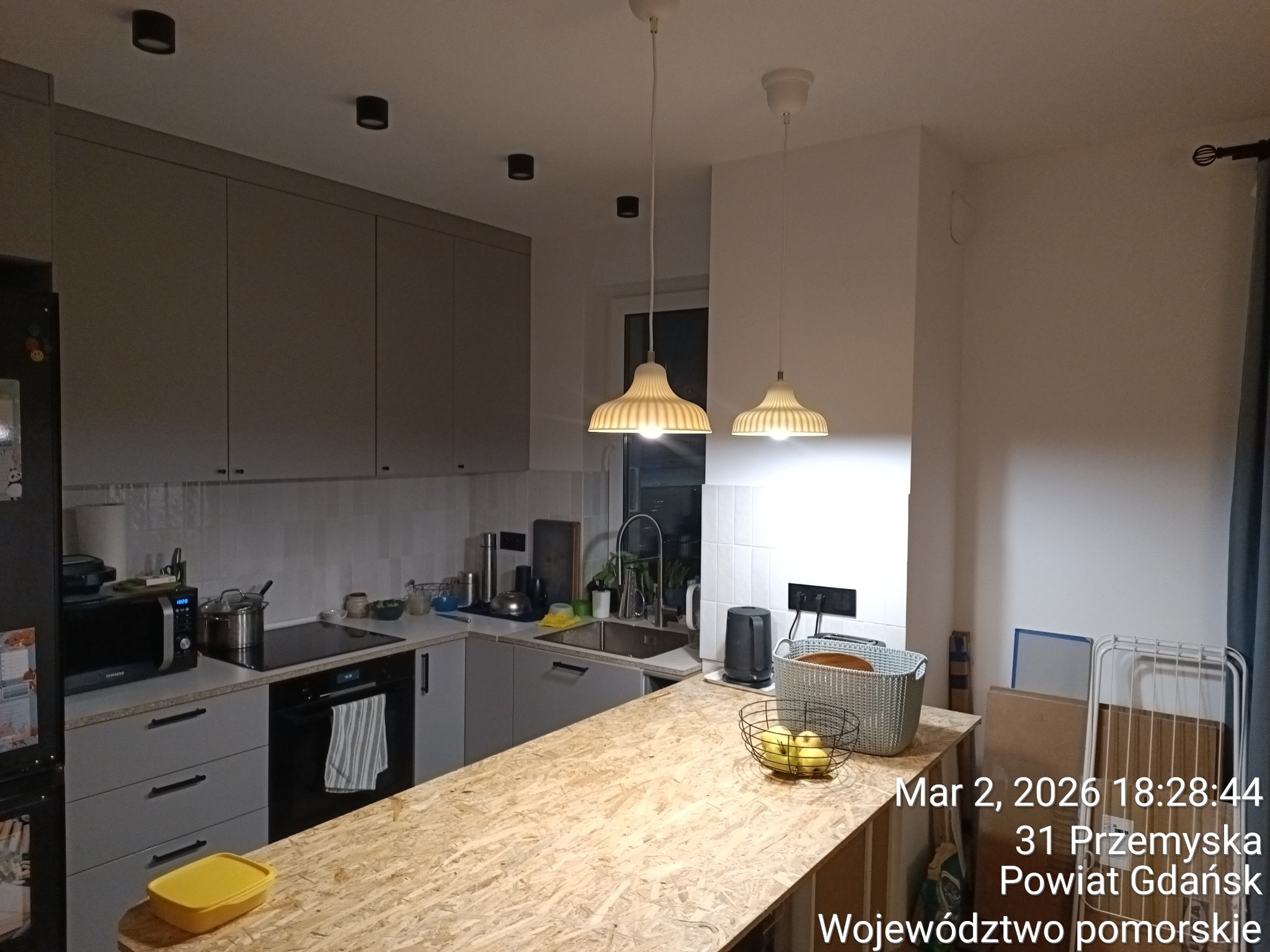 Jasna kuchnia z szarymi szafkami i blatem z płyty OSB. Dwie lampy wiszące nad blatem. Widoczne sprzęty AGD i akcesoria kuchenne. Minimalistyczny design.