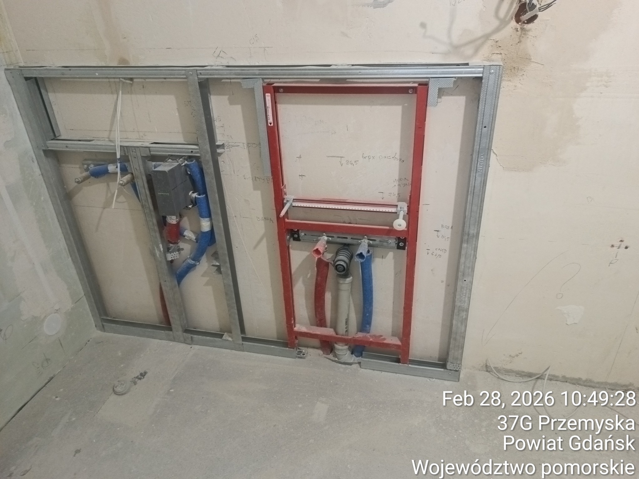 Stelaż podtynkowy WC z instalacją wodno-kanalizacyjną w trakcie montażu. Widoczne metalowe profile, rury doprowadzające wodę i odpływ. Surowe ściany w tle.