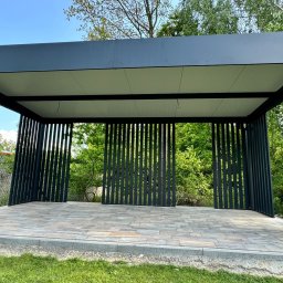 Anstal Rafał Babiarz - Nowoczesna, grafitowa pergola metalowa z pionowymi lamelami, wykończona kostką brukową, otoczona zielenią. Widok z zewnątrz, w tle drzewa.
