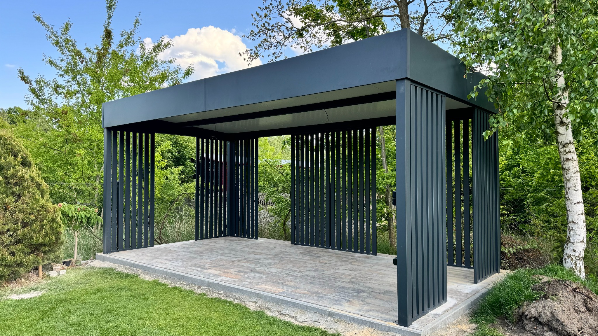 Nowoczesna, grafitowa pergola metalowa z pionowymi lamelami, osadzona na tarasie z kostki brukowej, otoczona zielenią ogrodu. Minimalistyczny design.
