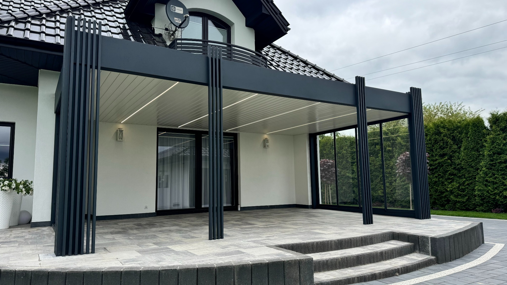 Nowoczesna, grafitowa pergola z oświetleniem LED, przylegająca do białego domu z czarnym dachem. Widoczne szklane panele boczne i taras z kostki brukowej.