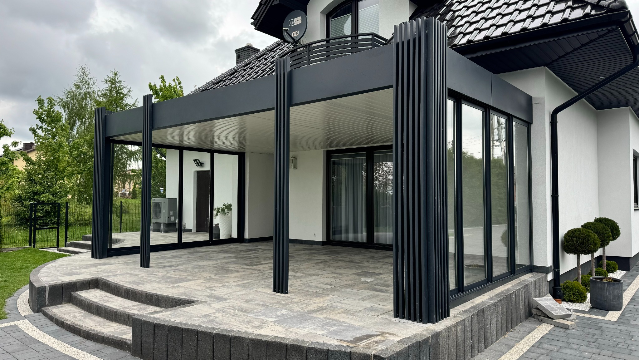 Nowoczesna, czarna pergola metalowa z przesuwnymi szklanymi ścianami, osłaniająca taras z kostki brukowej. Widok na dom z czarnym dachem i balkonem.