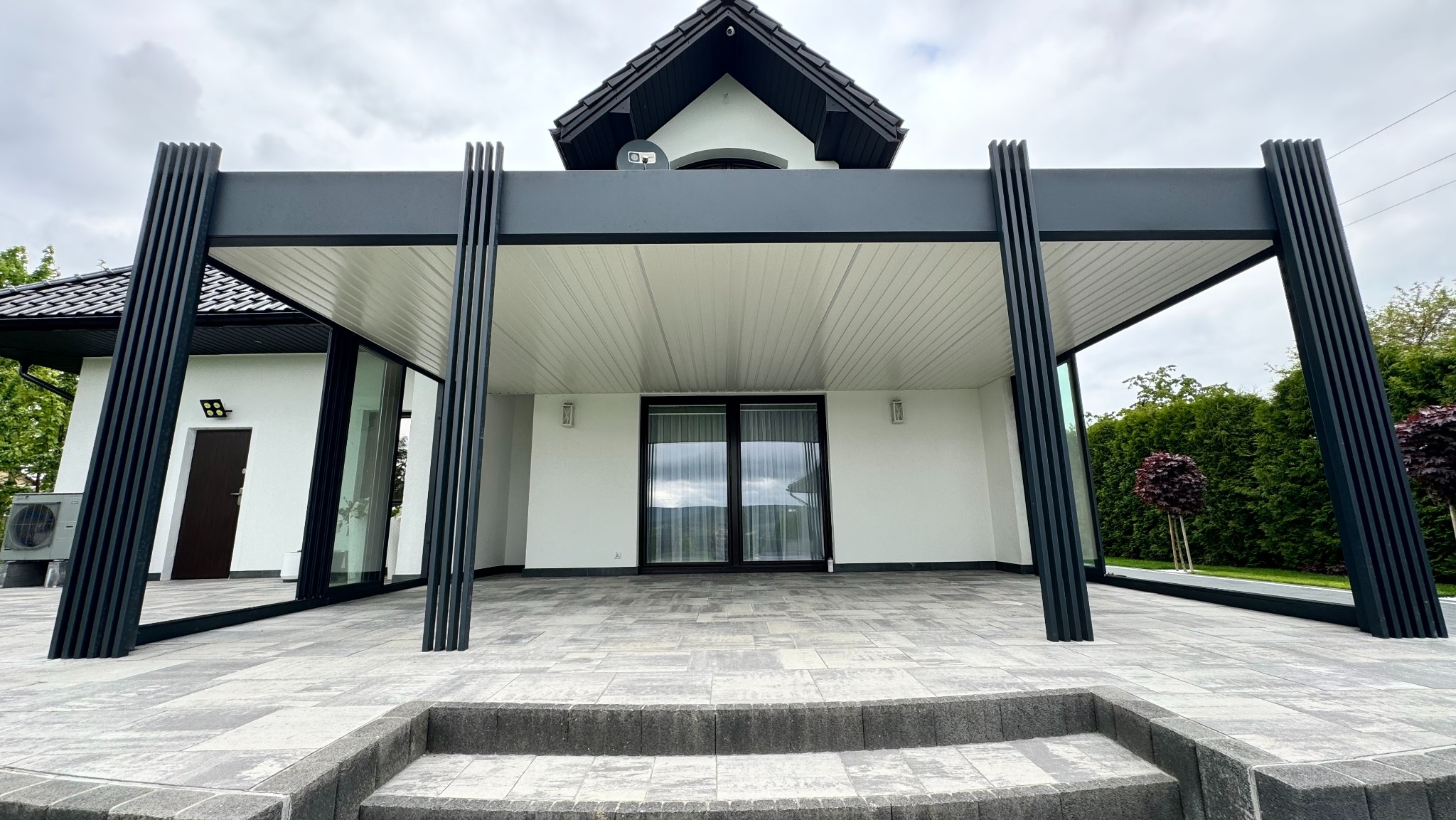 Nowoczesna, grafitowa pergola z lamelami, przylegająca do białego budynku z dużymi oknami. Kamienna posadzka tarasu z niskimi schodami. Minimalistyczny design.