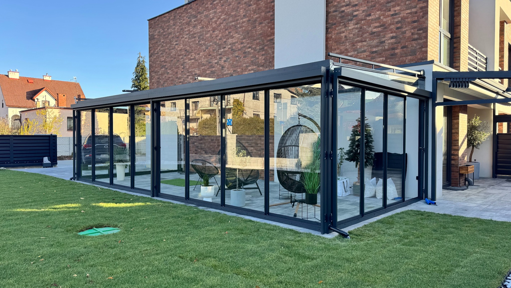 Nowoczesna zabudowa balkonu z przesuwnymi szklanymi panelami w grafitowej ramie, widok z zewnątrz na zadbany trawnik i elementy wyposażenia wnętrza.