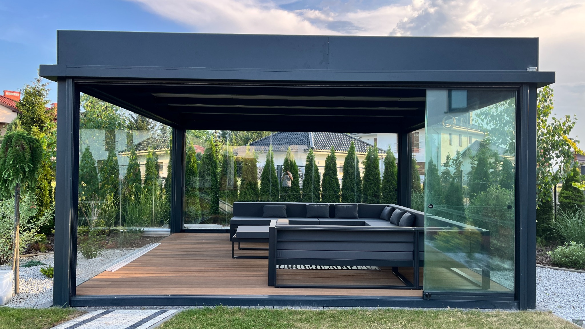 Nowoczesna, przeszklona pergola w antracytowym kolorze z drewnianym tarasem i meblami ogrodowymi. Widok na zadbany ogród z tujami i domem w tle.