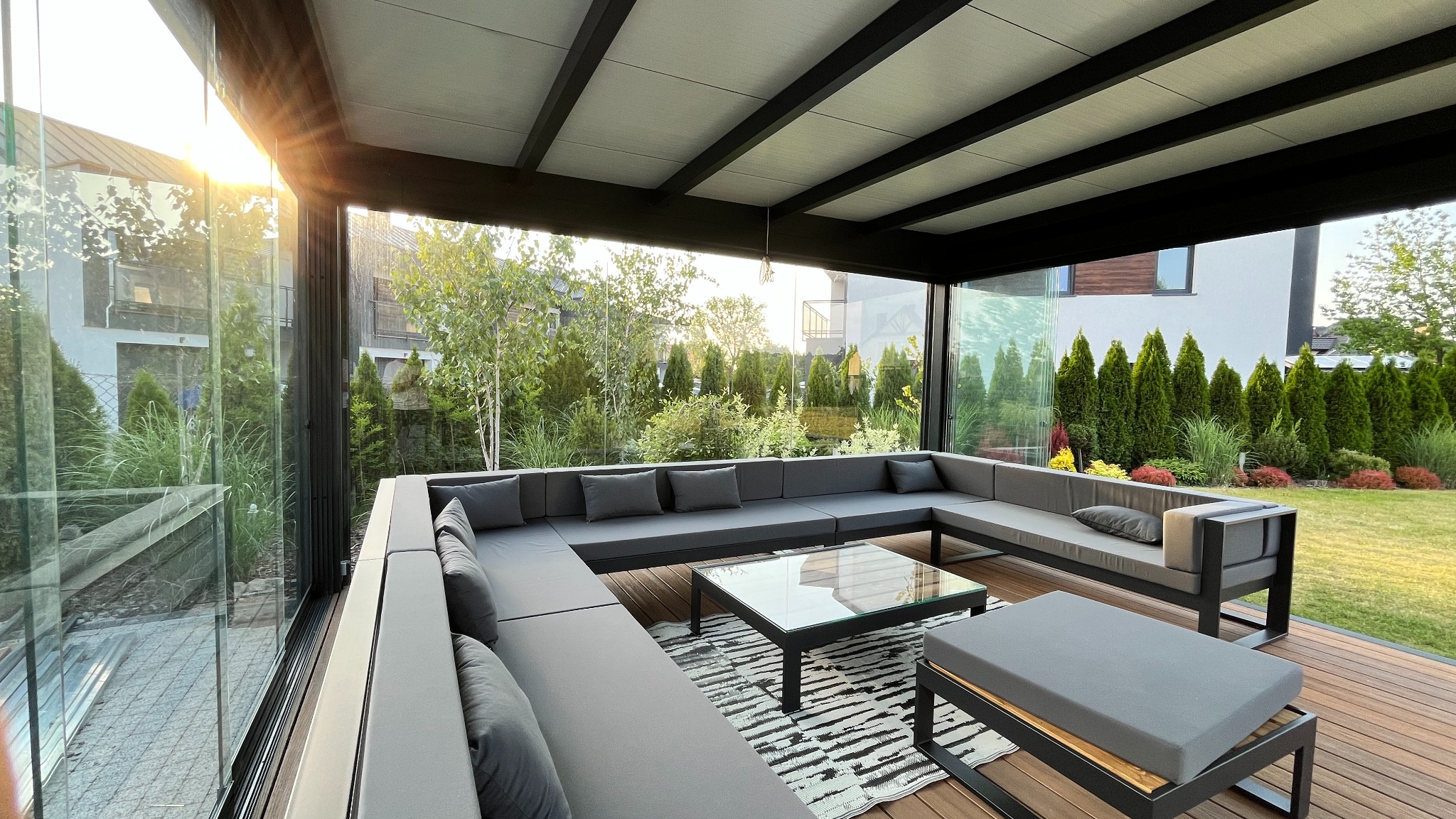 Nowoczesna zabudowa balkonu z metalową konstrukcją, szklanymi ścianami i komfortowym, szarym zestawem wypoczynkowym na drewnianym tarasie kompozytowym.