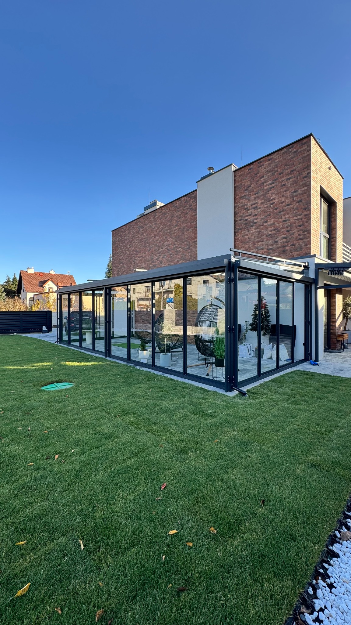 Nowoczesna, przeszklona pergola z metalową konstrukcją przylegająca do domu z ceglaną elewacją. Wewnątrz meble ogrodowe i dekoracje. Zadbany trawnik przed pergolą.