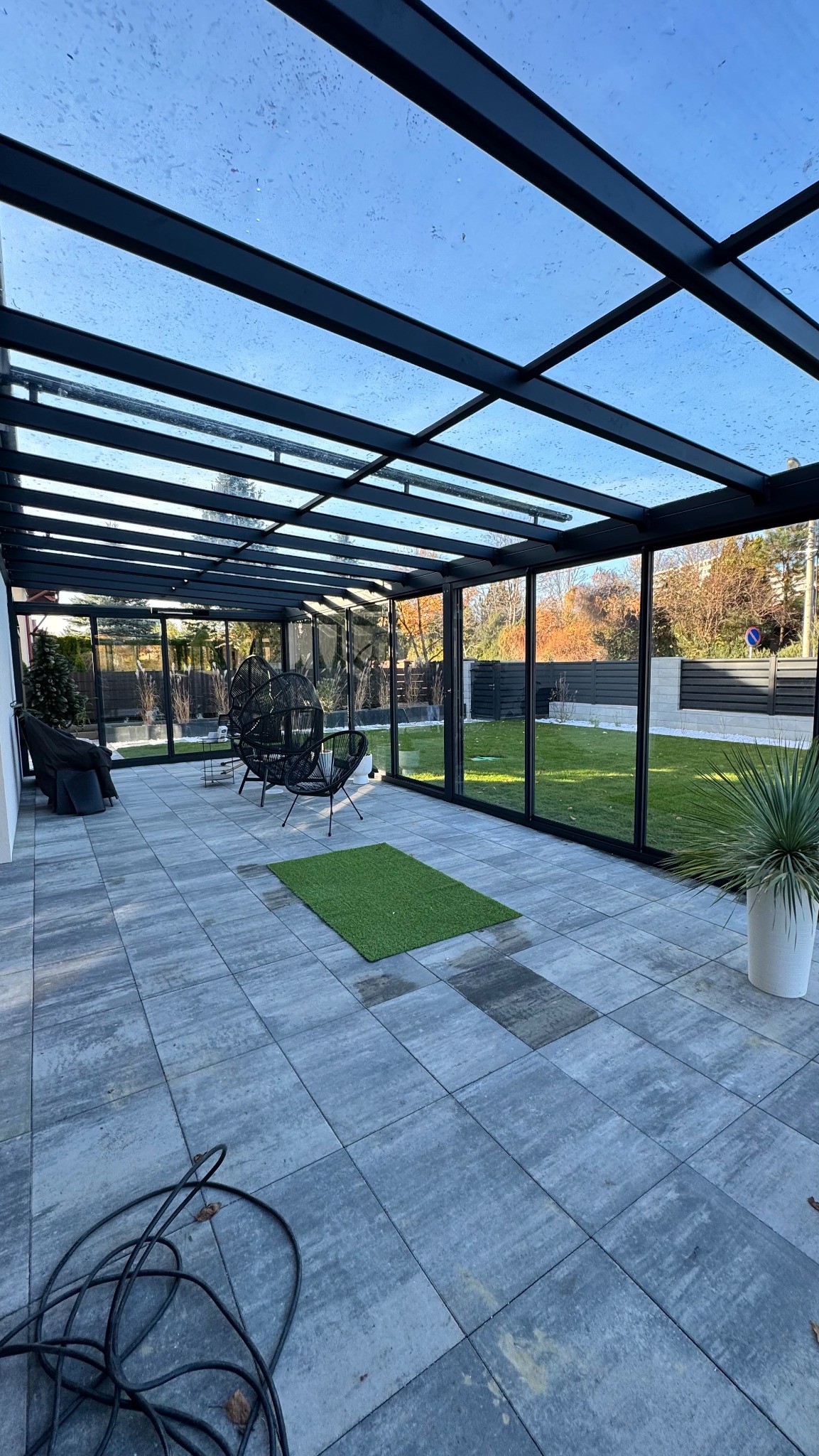 Nowoczesna zabudowa balkonu z metalową konstrukcją i szklanym dachem, widok na ogród. Na podłodze szare płyty i zielony dywanik. W tle czarne krzesła.