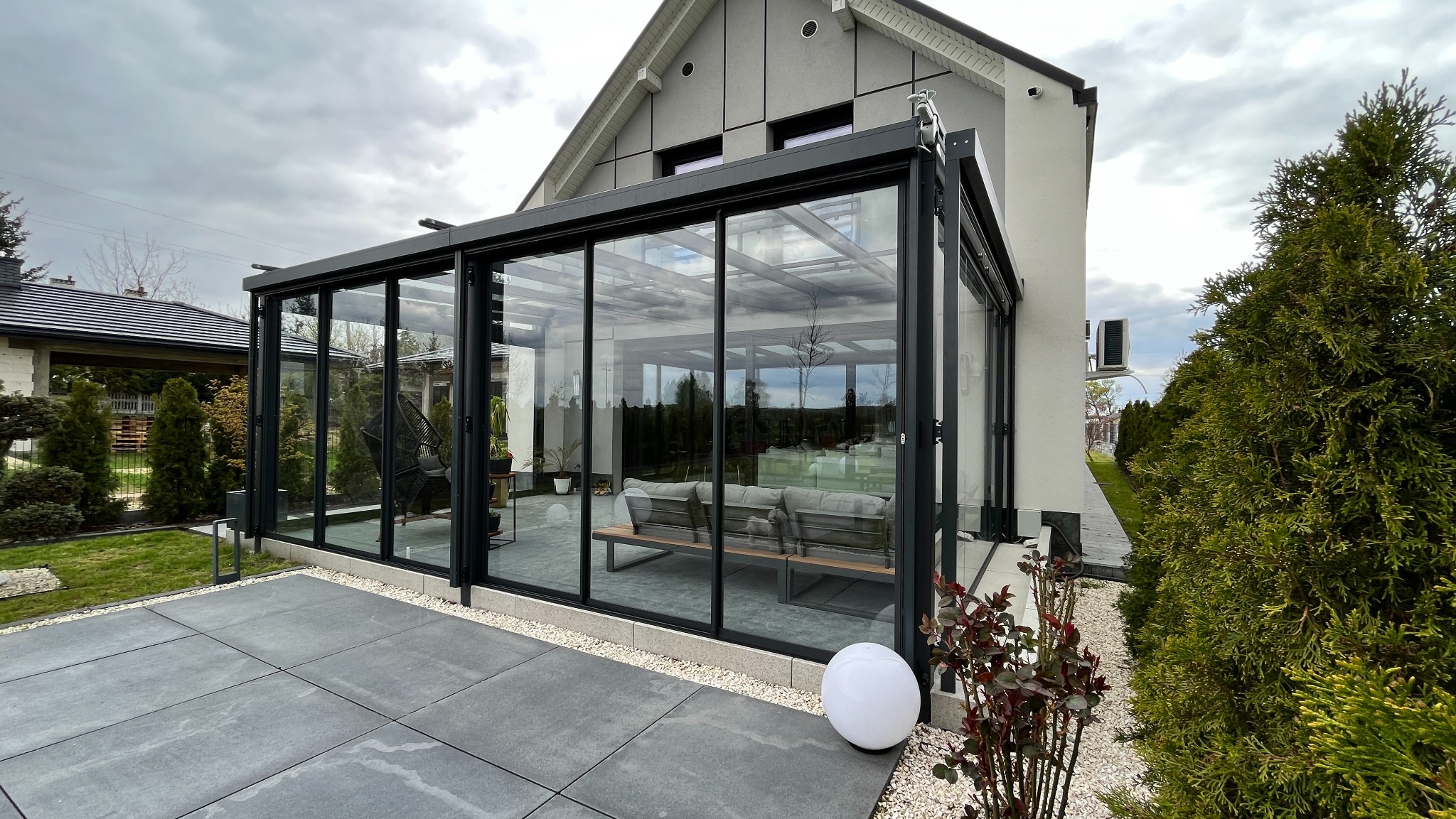 Nowoczesna zabudowa balkonu z czarnymi ramami i szklanymi panelami, widoczna z zewnątrz na tle szarej elewacji domu i zadbanego ogrodu. Wnętrze z meblami wypoczynkowymi.