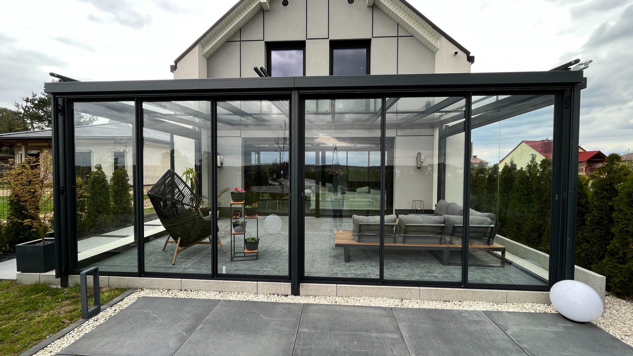 Szklana zabudowa tarasu z czarną ramą, widok z zewnątrz. Wewnątrz meble ogrodowe i dekoracje. Na zewnątrz szare płyty tarasowe i biała lampa.