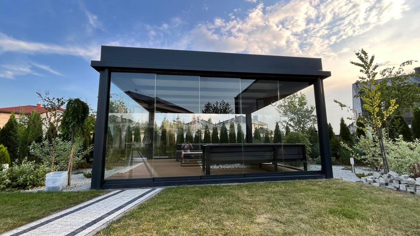 Nowoczesna, szklana zabudowa tarasu z czarną ramą, widok z zewnątrz na drewniany taras i meble ogrodowe. W tle zieleń i domy.