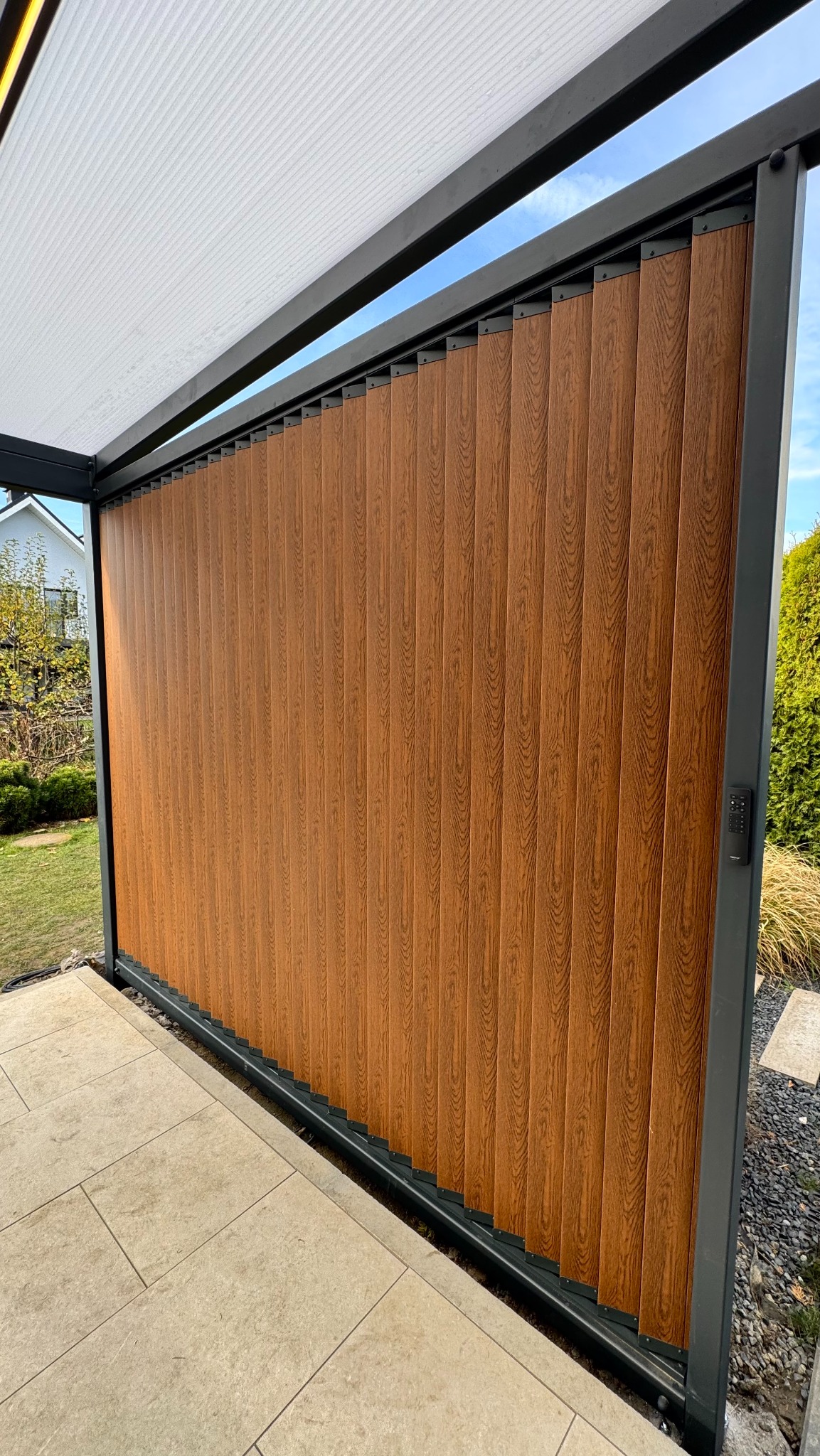 Nowoczesna pergola z drewnopodobnymi panelami i metalową konstrukcją, widok z zewnątrz na tarasie z jasną kostką. Minimalistyczny design, zamek cyfrowy na słupku.