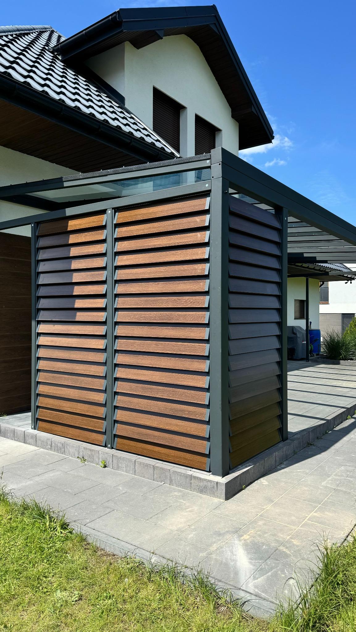 Nowoczesna pergola z drewnianymi żaluzjami i metalową konstrukcją, przylegająca do domu. Widoczne szare płyty chodnikowe i trawnik. Jasne, słoneczne oświetlenie.