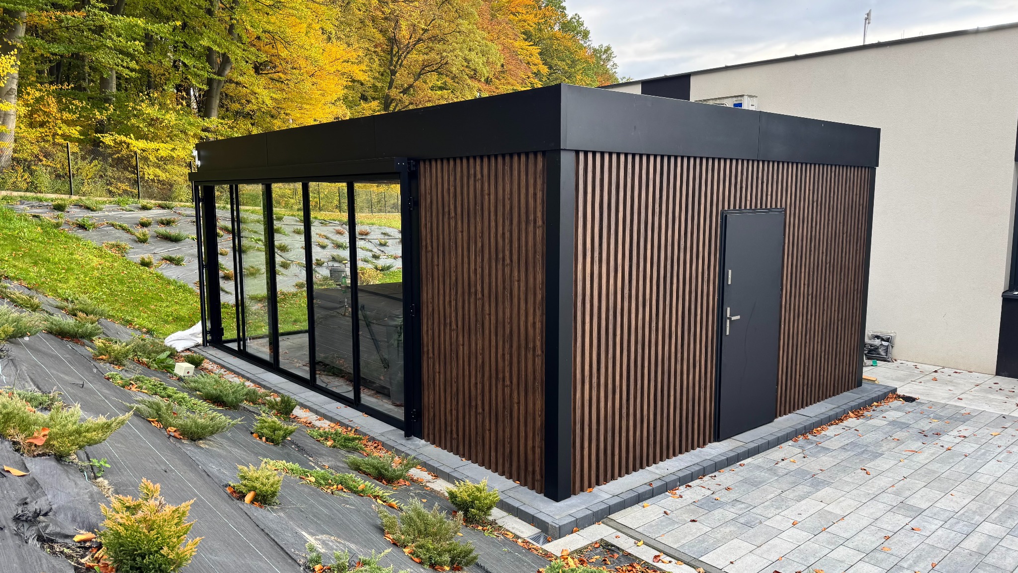 Nowoczesna, prostopadłościenna zabudowa z drewnianymi lamelami i czarną ramą okienną, otoczona zielenią i kostką brukową. Widok z zewnątrz, jesienna sceneria.