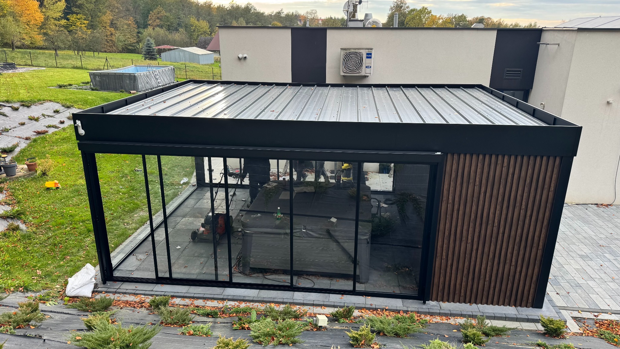 Nowoczesna, przeszklona pergola z metalową konstrukcją i drewnianymi panelami, z widocznym jacuzzi wewnątrz, otoczona zielenią i kostką brukową.