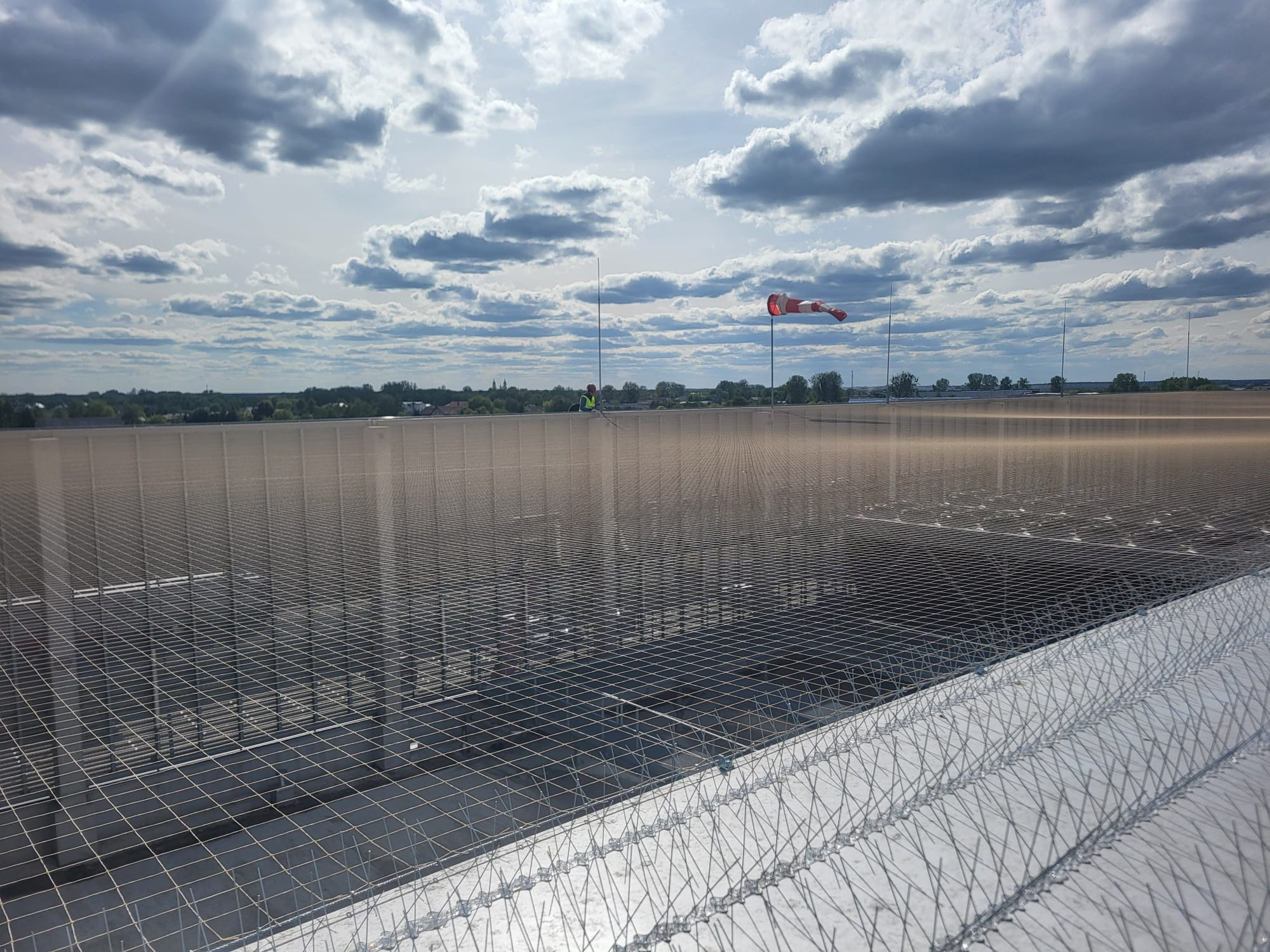 Siatka zabezpieczająca z kolcami na dachu budynku, widoczny w oddali pracownik w kamizelce, w tle pochmurne niebo i wskaźnik wiatru.