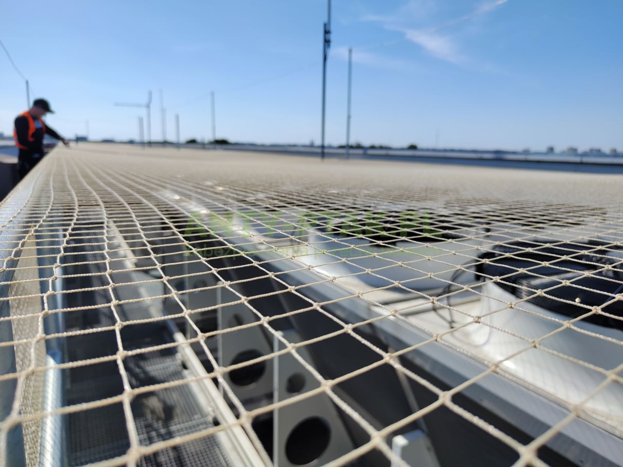 Siatka zabezpieczająca na dachu budynku, widok z bliska. W tle pracownik w kamizelce i panorama miasta pod błękitnym niebem. Zabezpieczenia na wysokości.