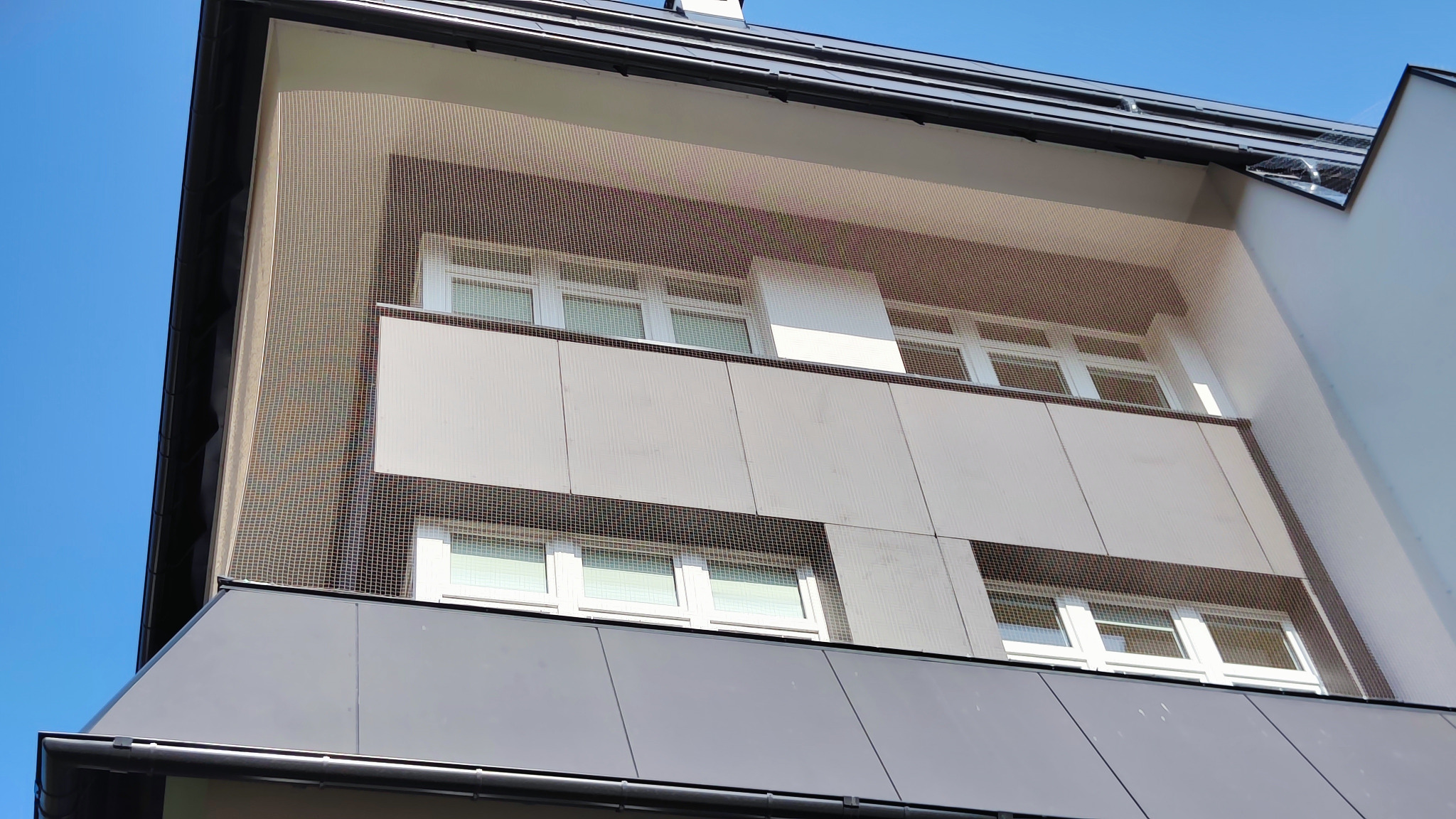 Siatka ochronna zamontowana na elewacji budynku, zabezpieczająca balkon i okna przed ptakami, widok z dołu na błękitne niebo.