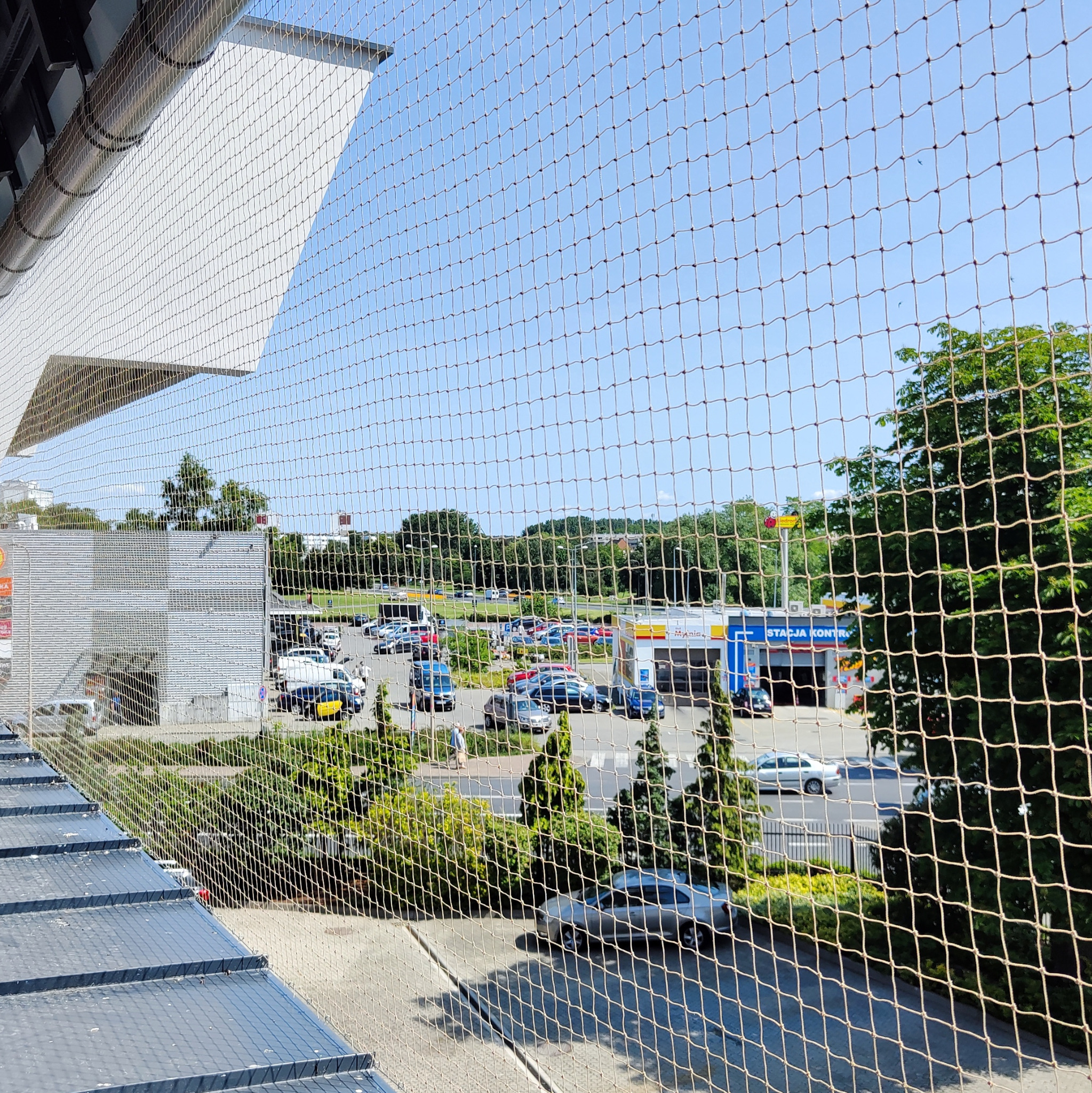 Zamontowana siatka ochronna na balkonie, zabezpieczająca przed wypadnięciem. Widok na parking i stację kontroli pojazdów w tle, przez oczka siatki.