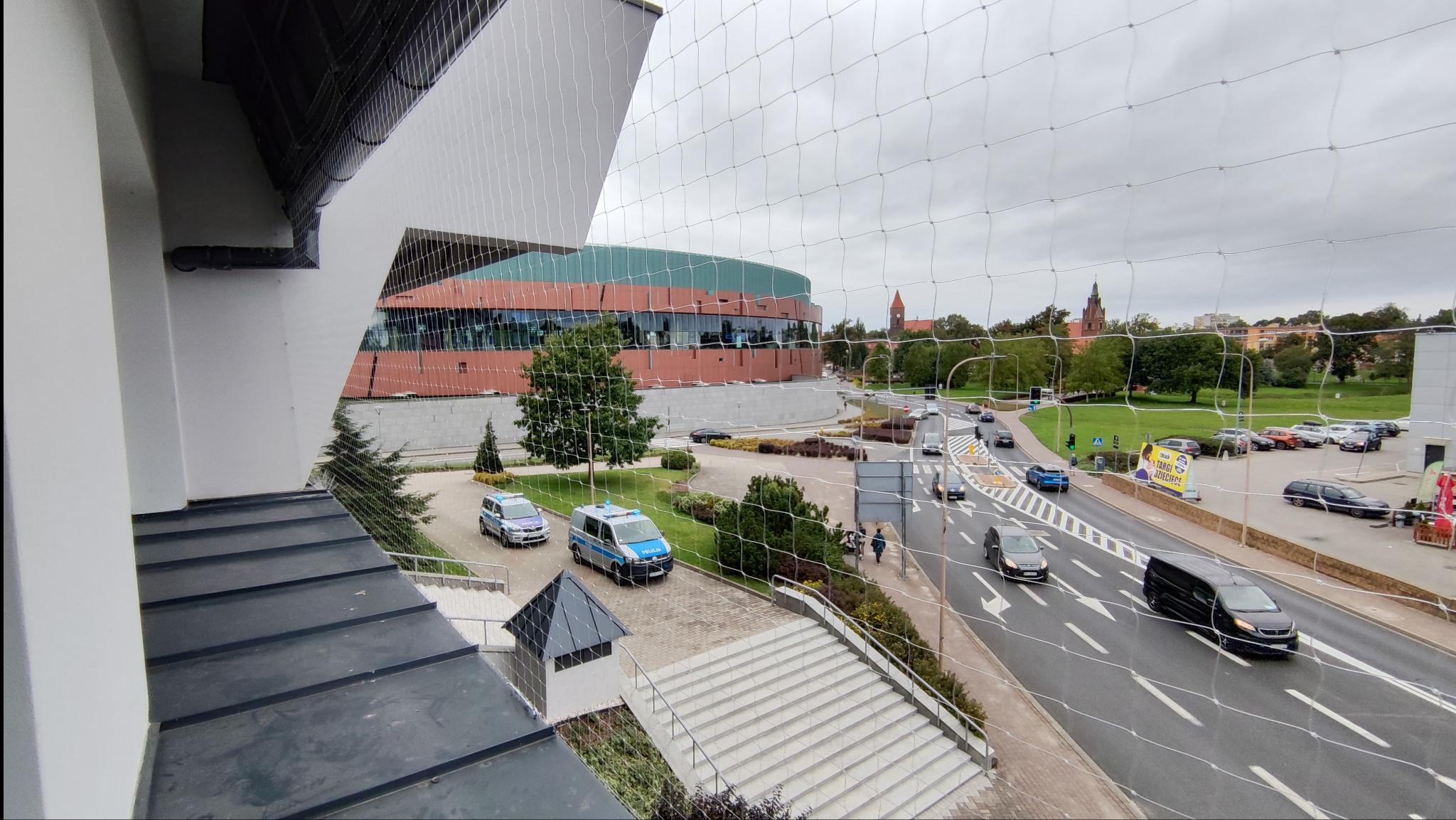 Siatka ochronna na balkonie z widokiem na miasto, zabezpieczająca przed ptakami. W tle budynek, ulica z samochodami i zieleń miejska.