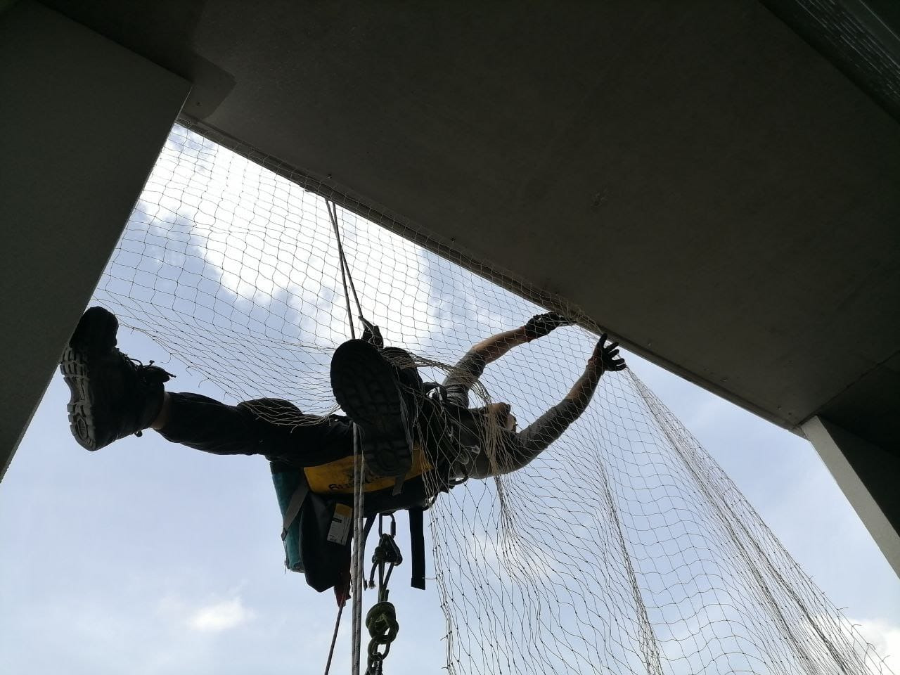 Pracownik zabezpieczony linami montuje siatkę ochronną pod konstrukcją mostu, widok z dołu na błękitne niebo.