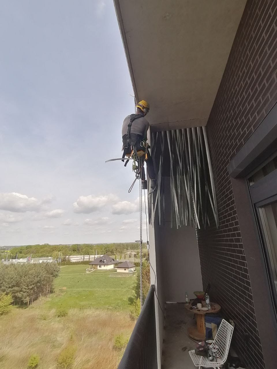 Pracownik na linie zabezpieczającej, w kasku, wykonuje prace na wysokości przy balkonie budynku z cegły. Widok z góry na krajobraz.