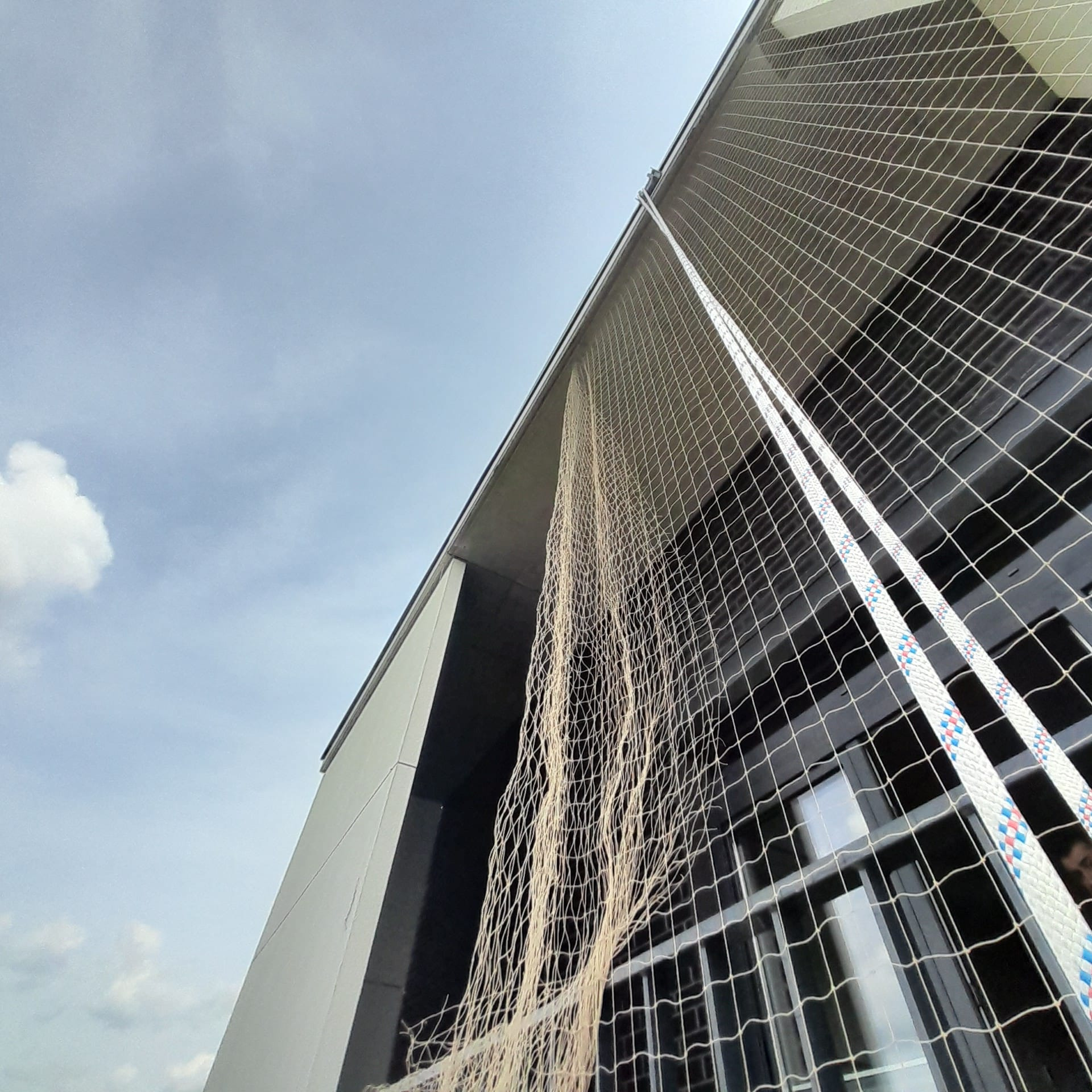 Siatka zabezpieczająca przed ptakami zamontowana na balkonie nowoczesnego budynku, widok z dołu, z fragmentem nieba w tle. Zabezpieczenie balkonu.