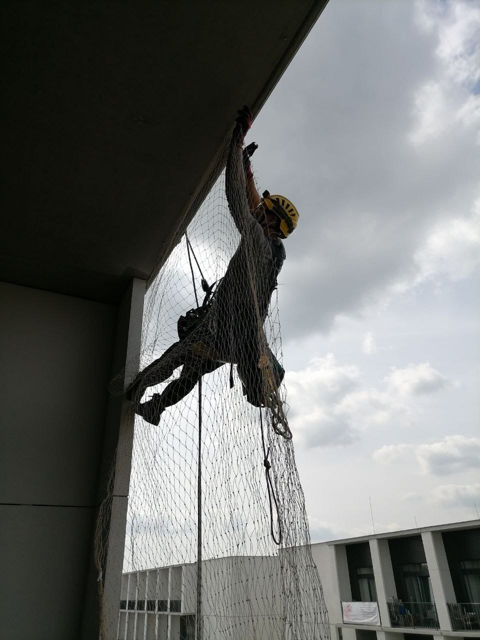 Pracownik w kasku, zabezpieczony linami, montuje siatkę ochronną na wysokości, na tle pochmurnego nieba i budynku. Prace wysokościowe.