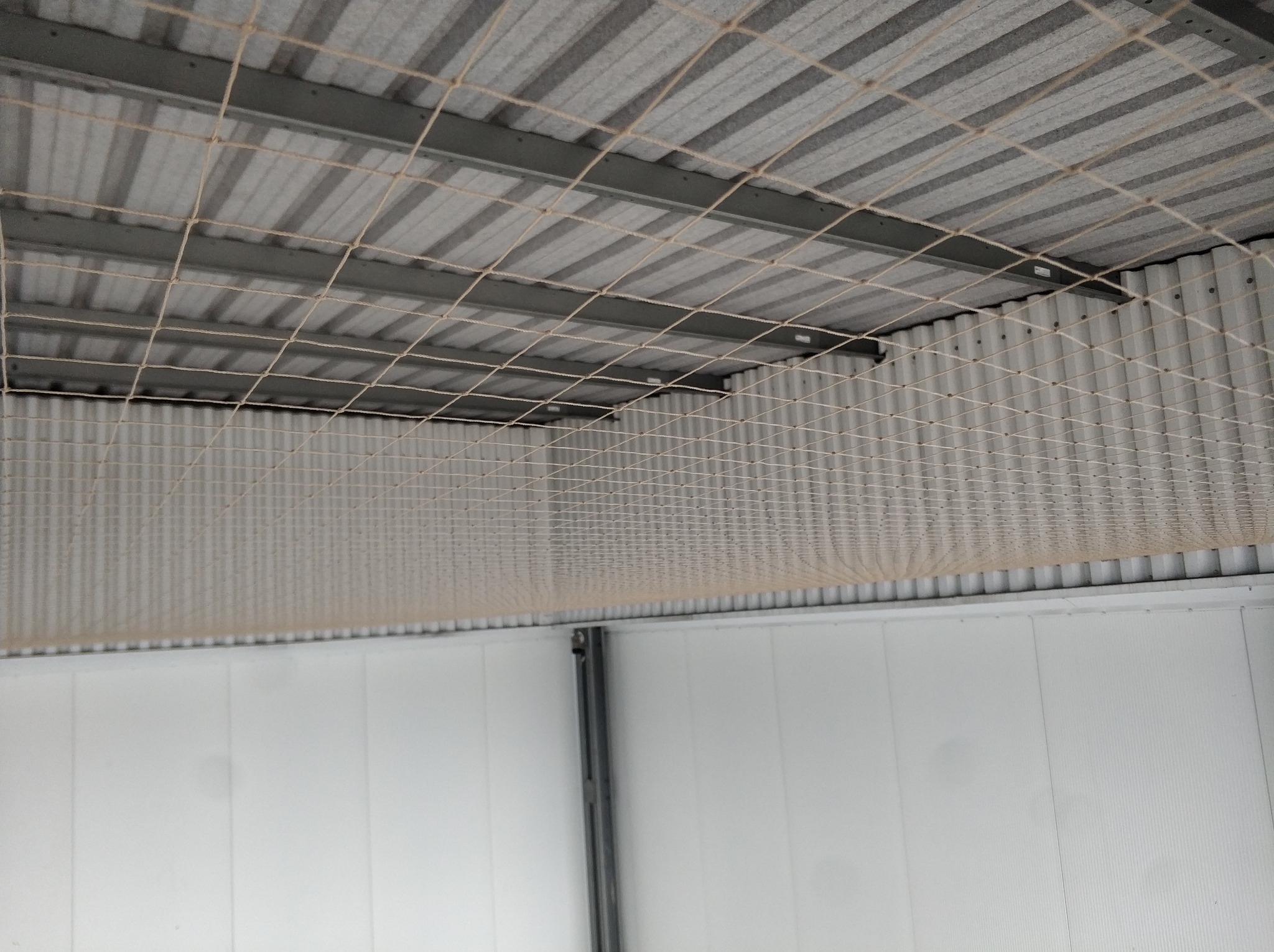Siatka zabezpieczająca zamontowana pod dachem z blachy falistej wewnątrz hali. Metalowa konstrukcja wsporcza. Białe ściany z paneli.