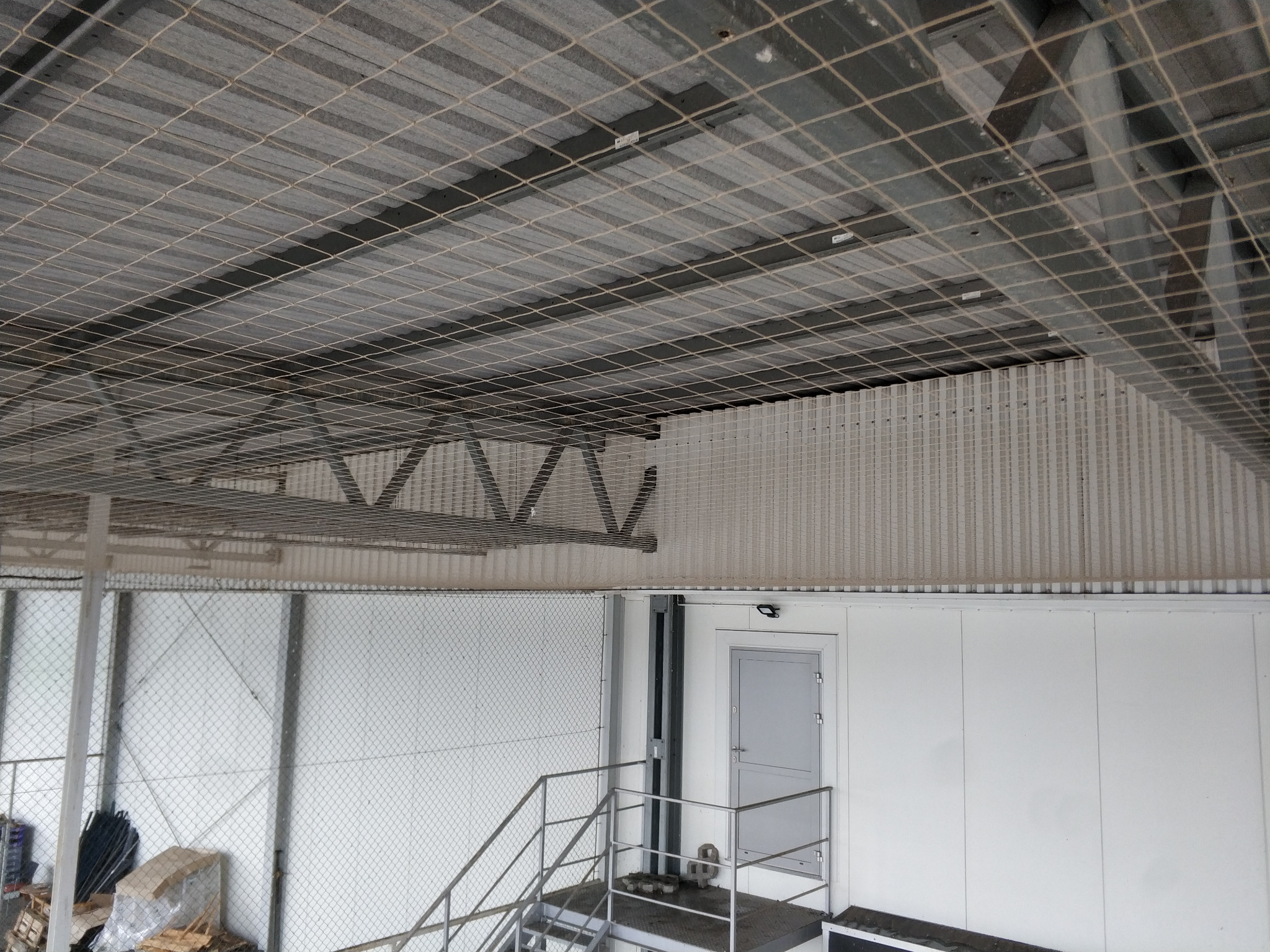 Siatka zabezpieczająca pod dachem hali magazynowej, widoczne schody i białe ściany. Zabezpieczenie przed spadającymi przedmiotami.
