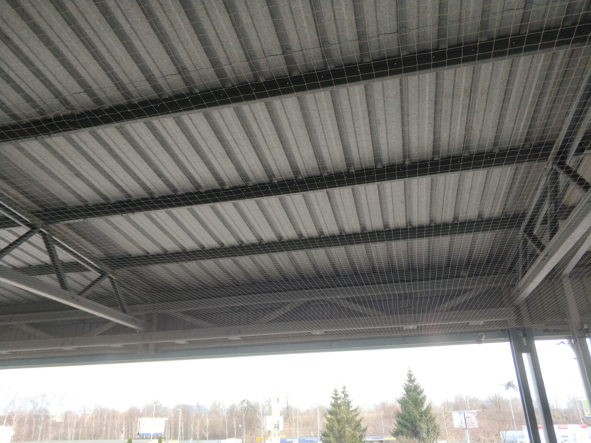 Siatka ochronna zamontowana pod metalowym zadaszeniem, zabezpieczająca przed ptakami. Widoczne elementy konstrukcyjne i otoczenie zewnętrzne.