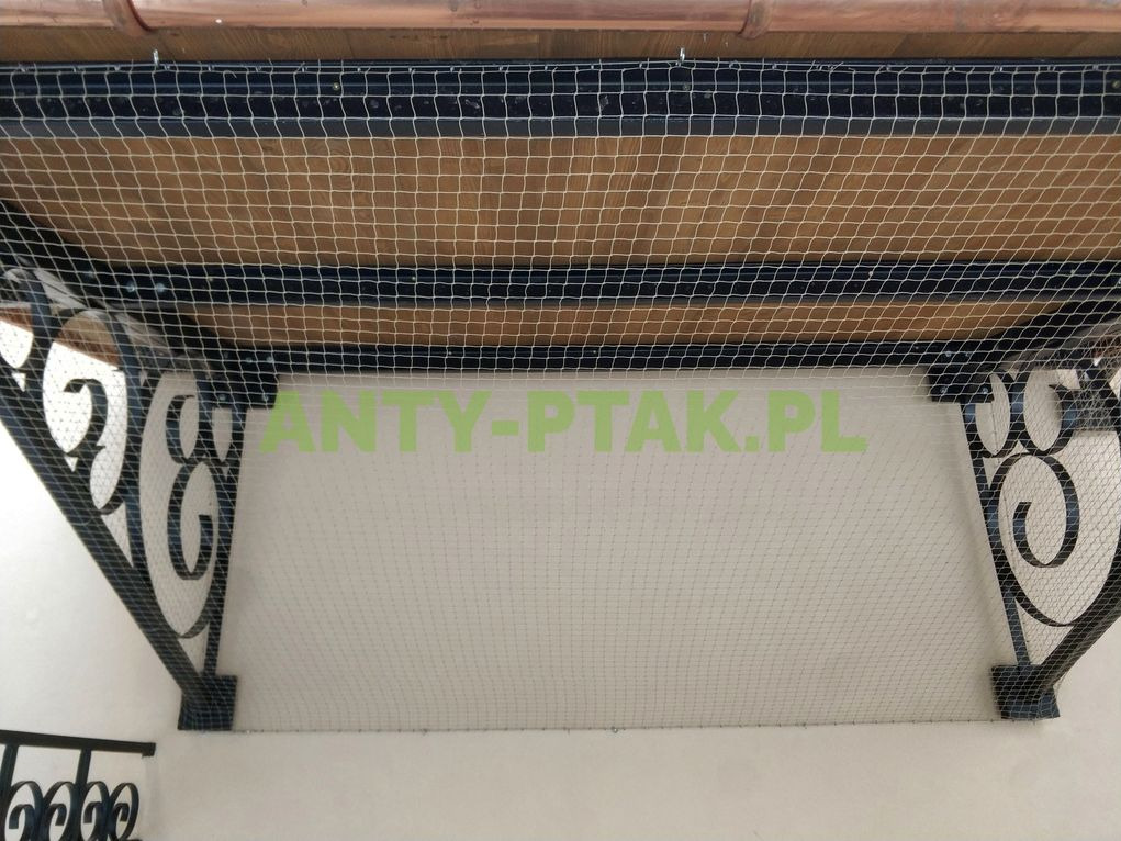 Zabezpieczenie balkonu siatką przeciw ptakom, widok od dołu. Metalowa konstrukcja wspornikowa z dekoracyjnymi elementami. Siatka naciągnięta na całej powierzchni.