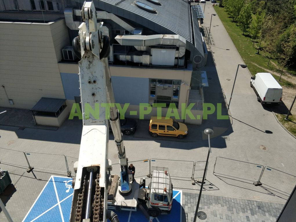 Widok z góry na podnośnik koszowy przy budynku z żółtym autem. Operator w koszu. Barierki zabezpieczające teren. Biały bus w oddali. Logo firmy na zdjęciu.