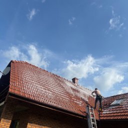 Skyline Malowanie i Renowacja Dachów Grzegorz Opyrchał - Pracownik w uprzęży czyści czerwoną dachówkę myjką ciśnieniową, tworząc wyraźną linię między umytą a brudną częścią dachu ceglanego domu, pod błękitnym niebem.