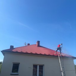 Skyline Malowanie i Renowacja Dachów Grzegorz Opyrchał - Pracownik w czerwonej koszulce i uprzęży bezpieczeństwa czyści czerwoną, falistą blachę dachową domu jednorodzinnego myjką ciśnieniową, stojąc na drabinie. Jasne ściany, błękitne niebo.