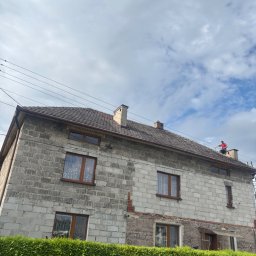 Skyline Malowanie i Renowacja Dachów Grzegorz Opyrchał - Pracownik w czerwonej koszulce na dachu dwupiętrowego domu z szarych pustaków, konserwujący brązowe pokrycie. Częściowo odsłonięta elewacja, zielony żywopłot.