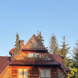 Skyline Malowanie i Renowacja Dachów Grzegorz Opyrchał - Pracownik w uprzęży na dachu drewnianego domu z ozdobnymi szczytami, używający sprzętu do czyszczenia lub malowania, oświetlony ciepłym słońcem.