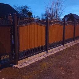 Denys Didych - Długi, dekoracyjny, brązowo-czarny płot z drewnianymi panelami i metalowymi zdobieniami, otaczający posesję. Widoczne elementy domu w tle, wieczorne oświetlenie.