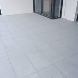Denys Didych - Jasnoszara, kwadratowa płytka na balkonie z widocznym śladem zabrudzenia w prawym dolnym rogu. W tle biała ściana i przesuwne drzwi balkonowe z czarną ramą.