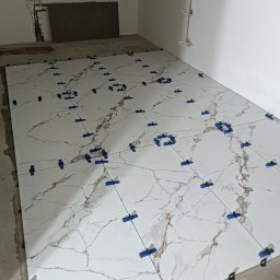 Denys Didych - Białe płytki imitujące marmur, ułożone na podłodze z użyciem niebieskich klipsów poziomujących. Widoczne kable elektryczne w ścianie i surowy stan wykończenia.