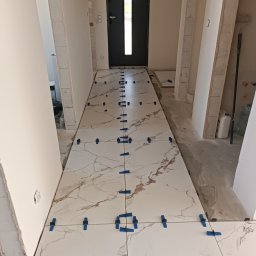 Denys Didych - Białe płytki imitujące marmur, ułożone w korytarzu, z niebieskimi klipsami poziomującymi. Widoczny fragment drzwi wejściowych i surowy stan ścian.