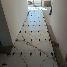 Denys Didych - Białe płytki imitujące marmur, ułożone w korytarzu, z niebieskimi klipsami poziomującymi. Widok na zewnątrz przez otwarte drzwi.