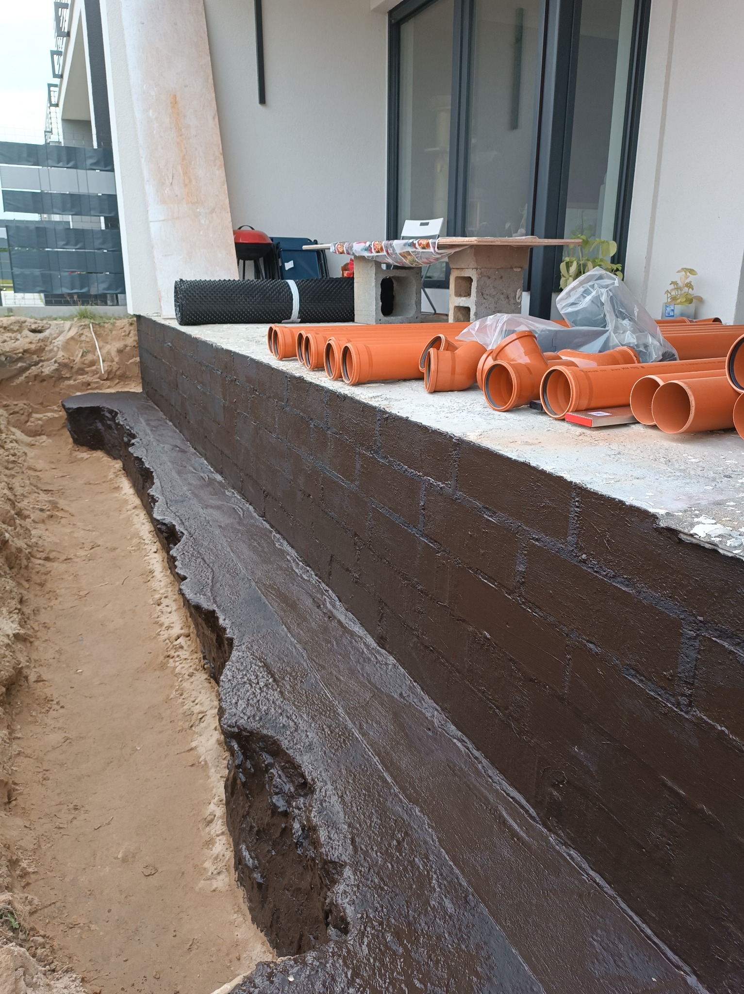 Zabezpieczona hydroizolacją ściana fundamentowa z wykopem, przygotowana do dalszych prac. Na górze rury kanalizacyjne i materiały budowlane.