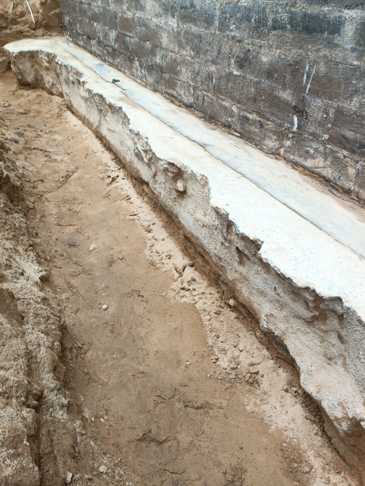 Wykonany fundament pod budynek z widoczną izolacją poziomą, otoczony wykopem z piaskiem. Ściana budynku z cegły w tle.